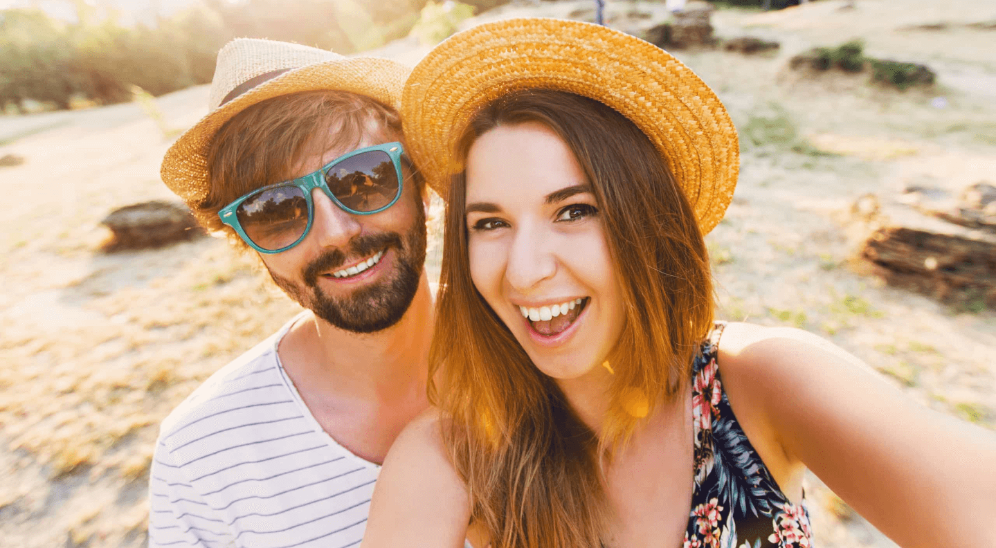Happy young couple wearing summer hats and sunglasses, smiling outdoors in a sunny park setting