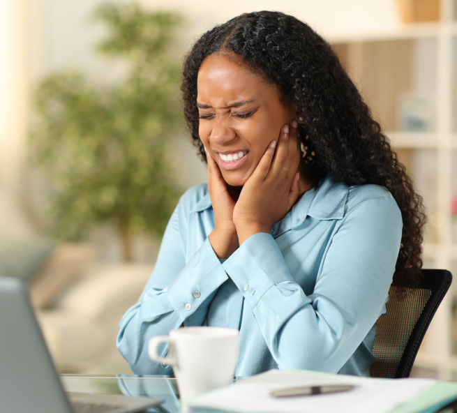 Woman experiencing tooth pain at desk, touching her cheek while sitting at workspace.