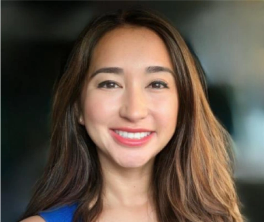 Professional headshot of smiling woman with long brown hair wearing blue top in dental office setting.